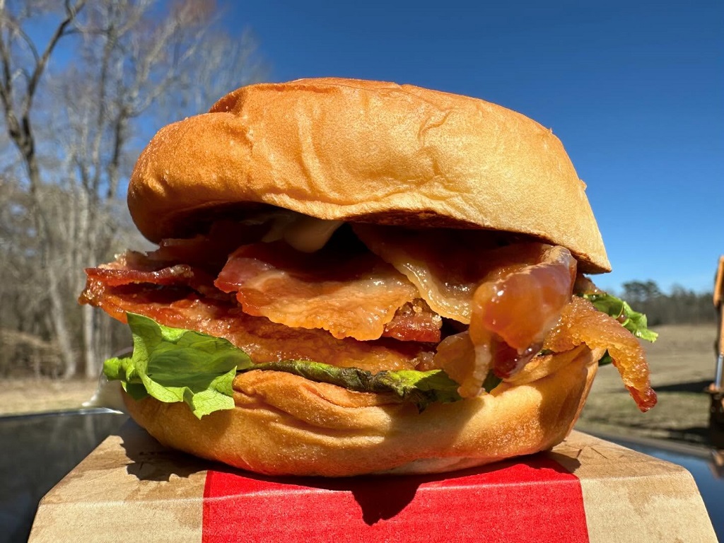 Arby’s breakfast sandwich with crispy bacon, lettuce, and sauce under clear skies