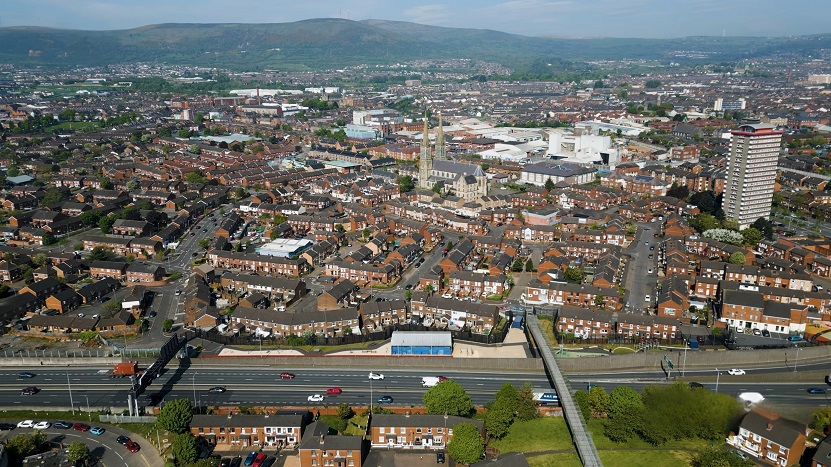 Aerial view of Belfast featuring dense rows of red-brick houses, a prominent church, high-rise buildings, and green hills in the background