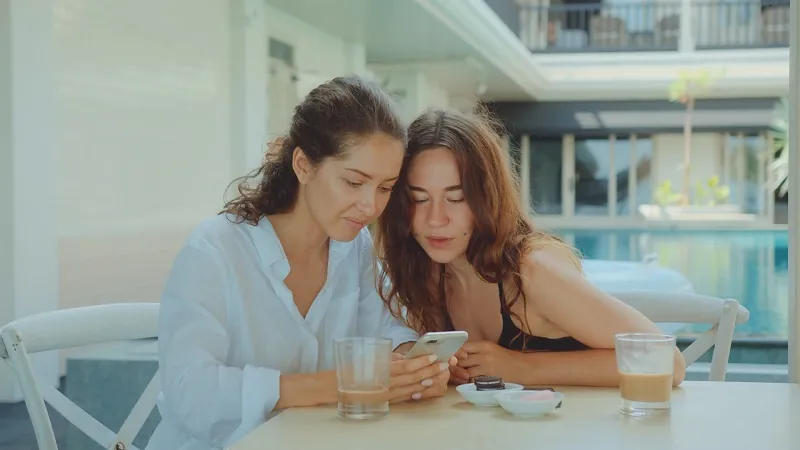 Mother and daughter sitting together at a table, smiling while looking at a phone