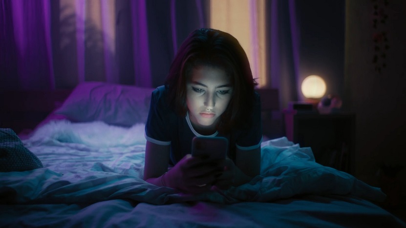 Teenage girl lying on a bed in a dark room, illuminated by the light of her smartphone screen