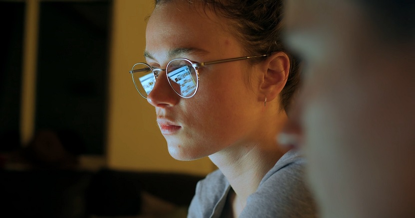 Close-up of a young woman wearing glasses, with a screen reflected in the lenses as she looks at a digital device in low light