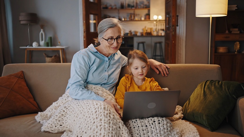 An older woman and a young girl sitting on a couch under a blanket, looking at a laptop together