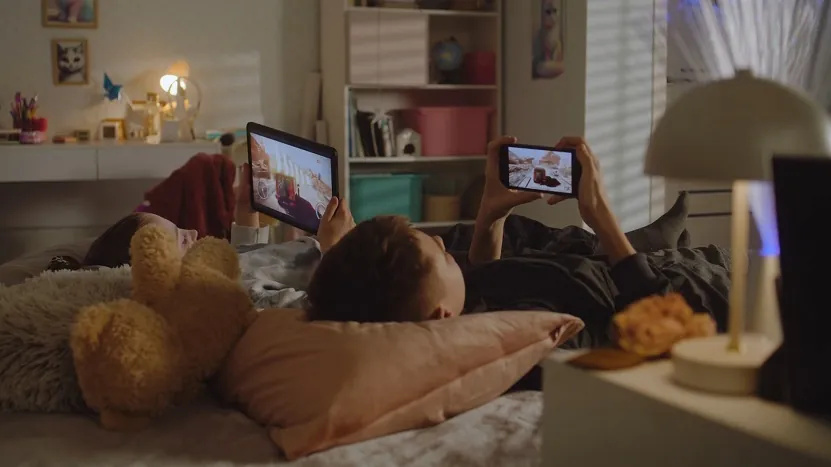 Two children lying on a bed watching videos on a tablet and a smartphone in a dimly lit bedroom
