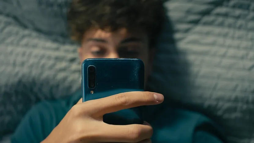 Teenager lying in bed, focused on a smartphone screen in a dimly lit room