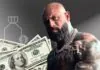 Tattooed, muscular man wearing chains in front of U.S. $100 bills and a money bag icon on a gray background