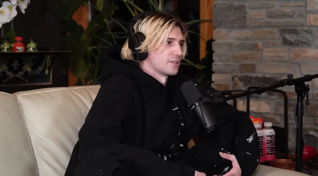 xQc speaking into a microphone during a podcast interview, wearing a black hoodie and headphones