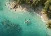 Aerial view of a tropical beach with clear turquoise water, palm trees, and an airplane flying low above the shoreline