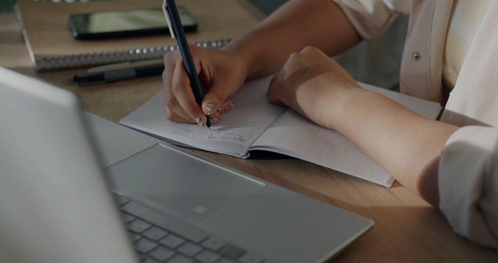 Person writing notes by hand beside an open laptop